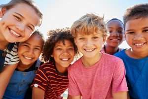 A group of 6 children leaning into the camera and smiling on a sunny day.