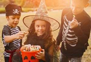 3 children in Halloween costumes are taking candy from an orange bucket one is holding