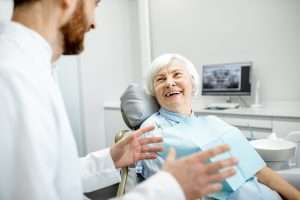 A senior with white hair is sitting in a dentist's chair and smiling at a person in a white coat who is gesturing.