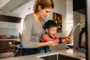 An adult in a gray shirt holds up a child so they can wash their hands from the kitchen faucet.